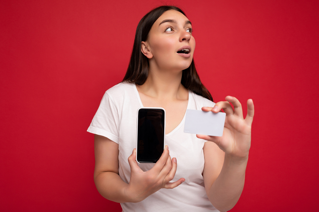 Photo of Beautiful Surprised Young Brunette Woman Wearing White T-Shirt Isolated on Red Background Holding Credit Card and Mobile Phone with Empty Display for Cutout Looking to the Side